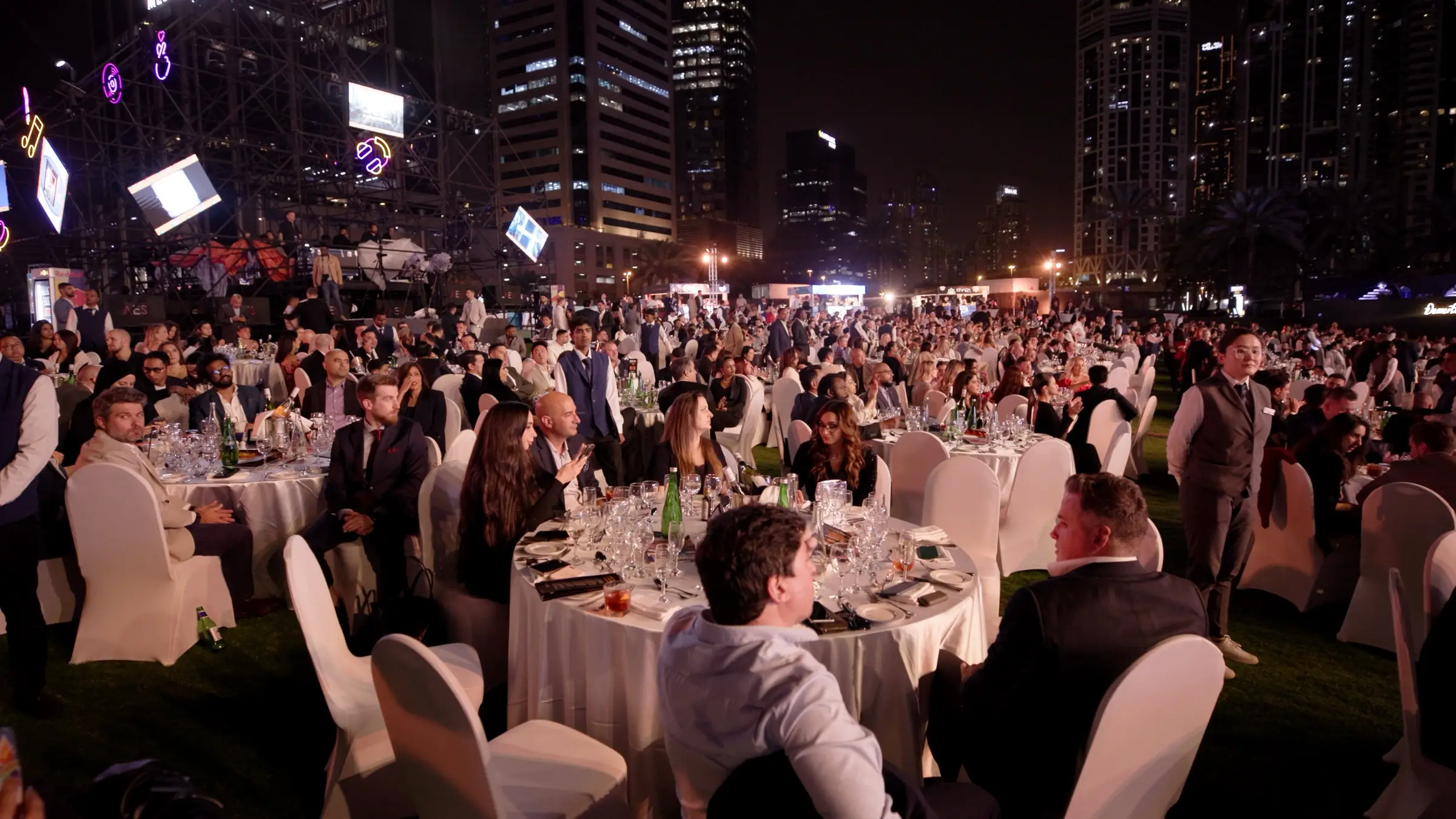 A large crowd standing at an outdoor awards ceremony lit in pink and purple