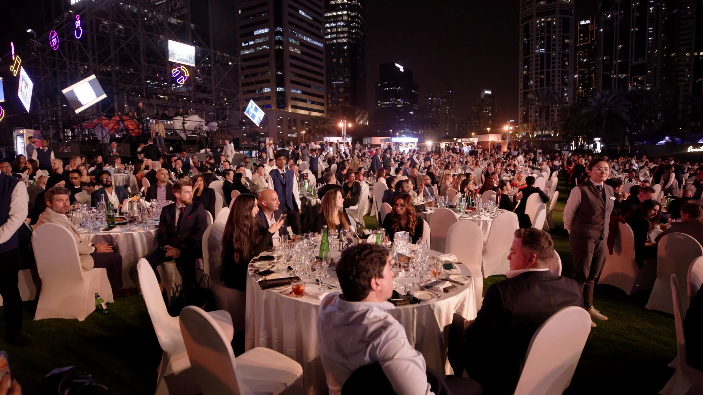 A large crowd standing at an outdoor awards ceremony lit in pink and purple