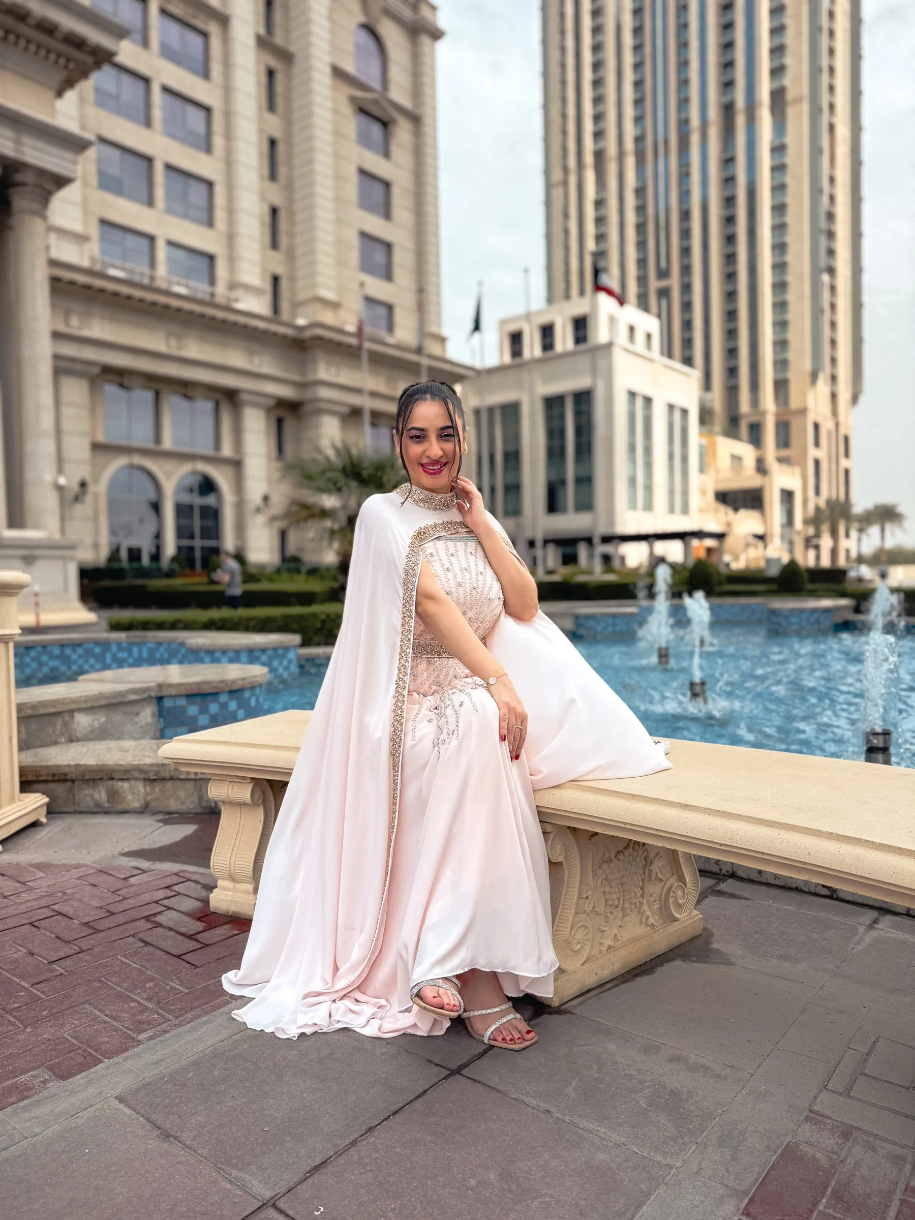 A bride in a white gown seated beside an ornamental fountain in a palace courtyard