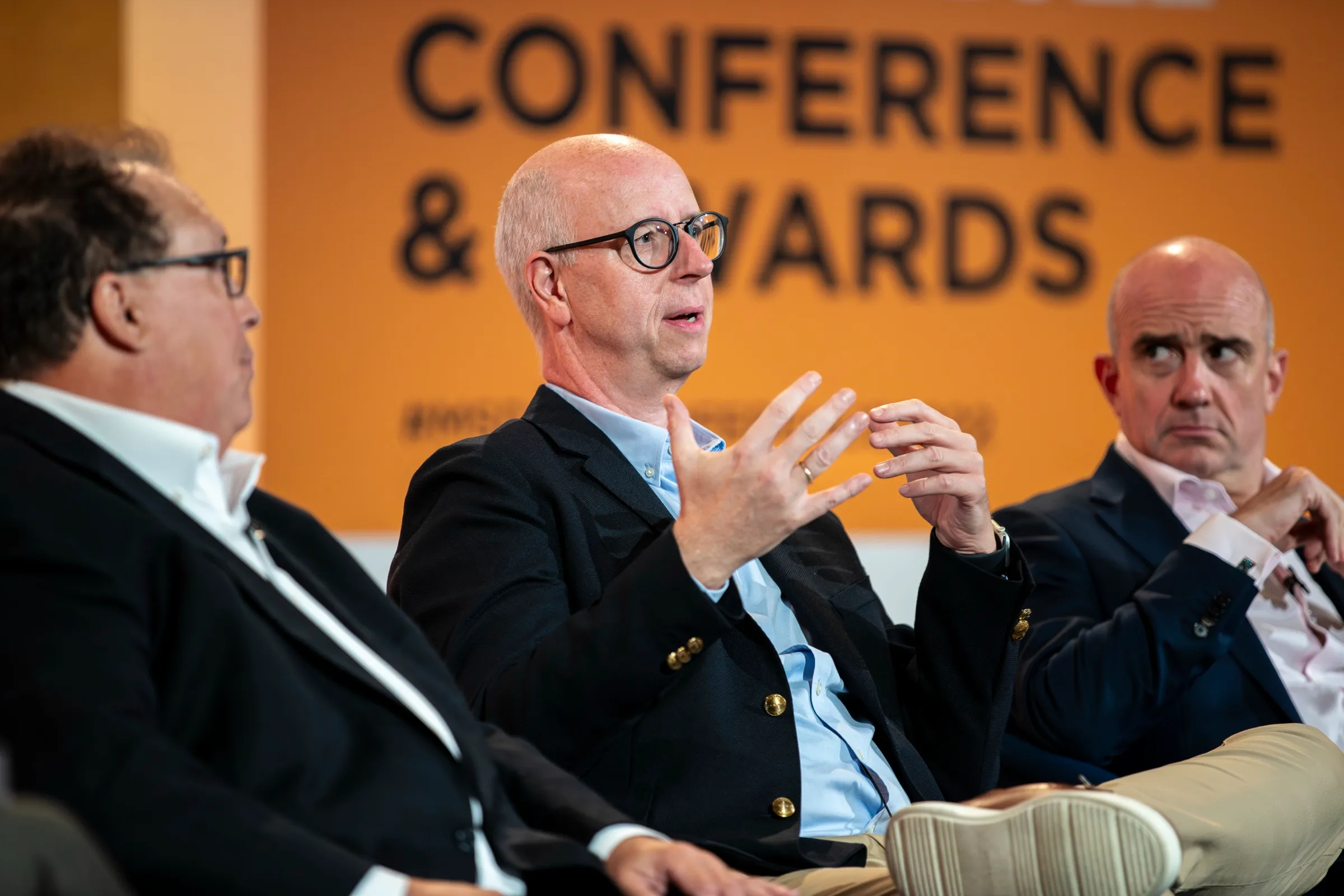 Three panellists in discussion on stage at a conference beneath a Conference and Awards backdrop