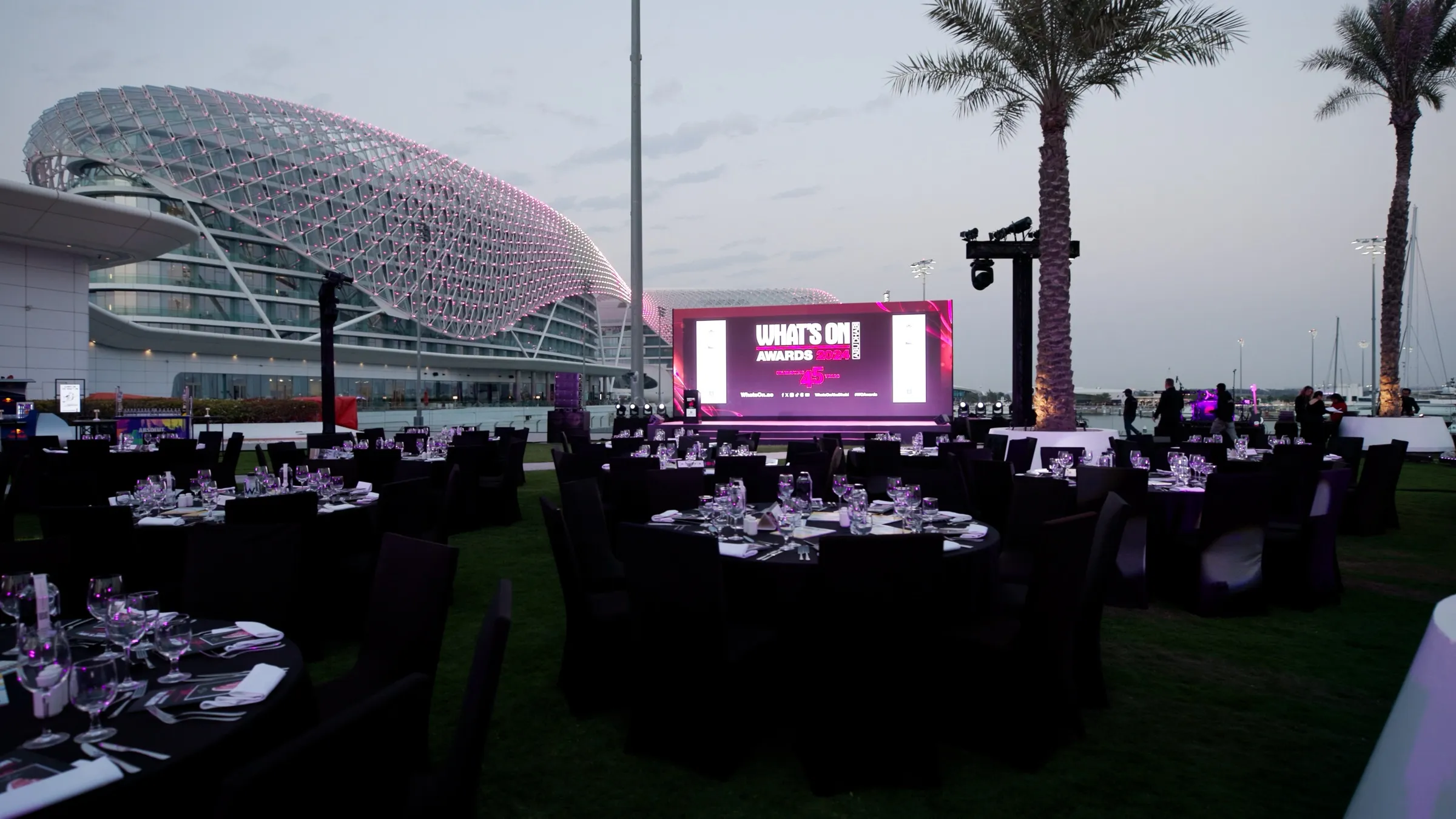 An outdoor gala dinner set on a waterfront terrace at dusk with a large stage screen