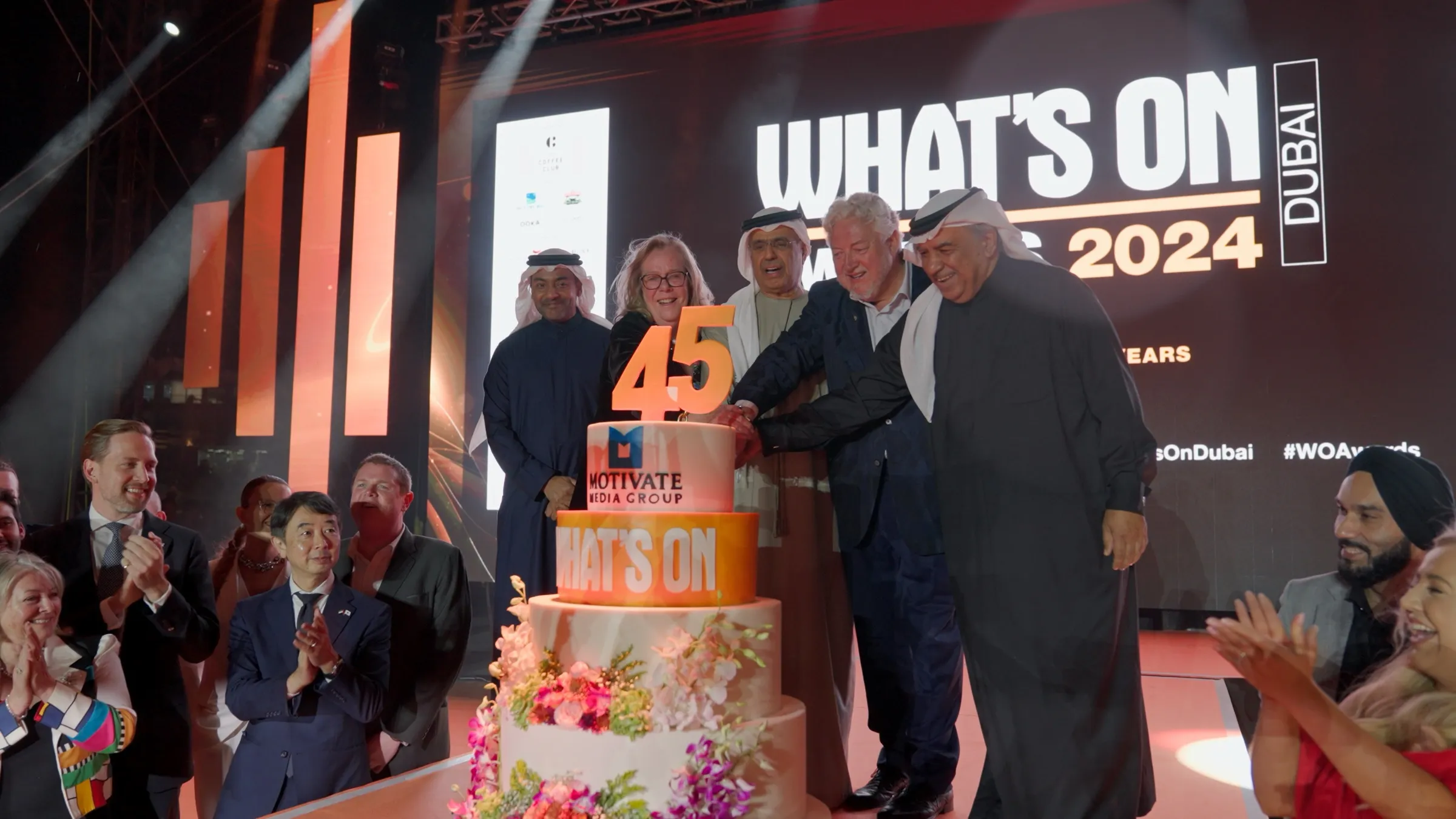Five guests of honour cutting a tiered anniversary cake on stage as the audience applauds