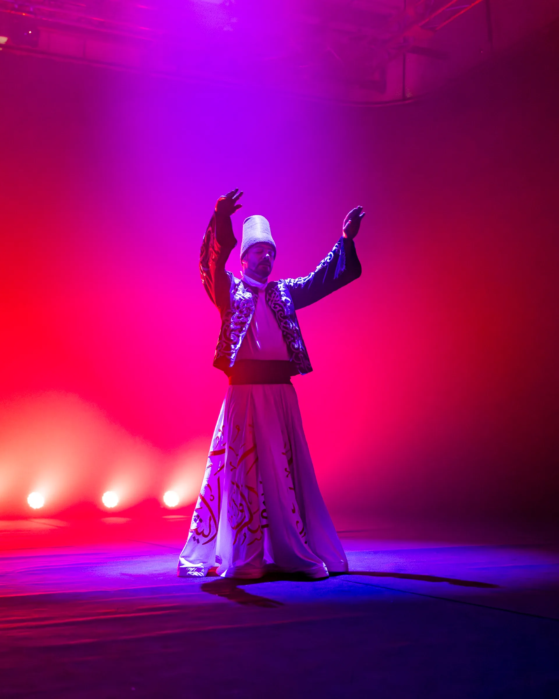 A whirling dervish performer with arms raised, backlit in magenta and pink